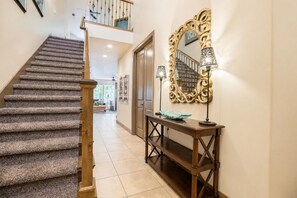 Un escalier dont les marches sont recouvertes de moquette, menant à un deuxième étage, une console avec un miroir et des lampes, et une vue sur l’espace de vie.