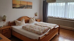 Een slaapkamer met een houten bed, bloemenbeddengoed, een nachtkastje met lamp, een wandplank en een raam met gordijnen.