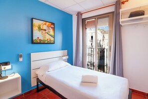 A hotel room with a bed, a nightstand, a painting, a window with a view of buildings, and a balcony. - Hostal Forum Tarragona (Tarragona)
