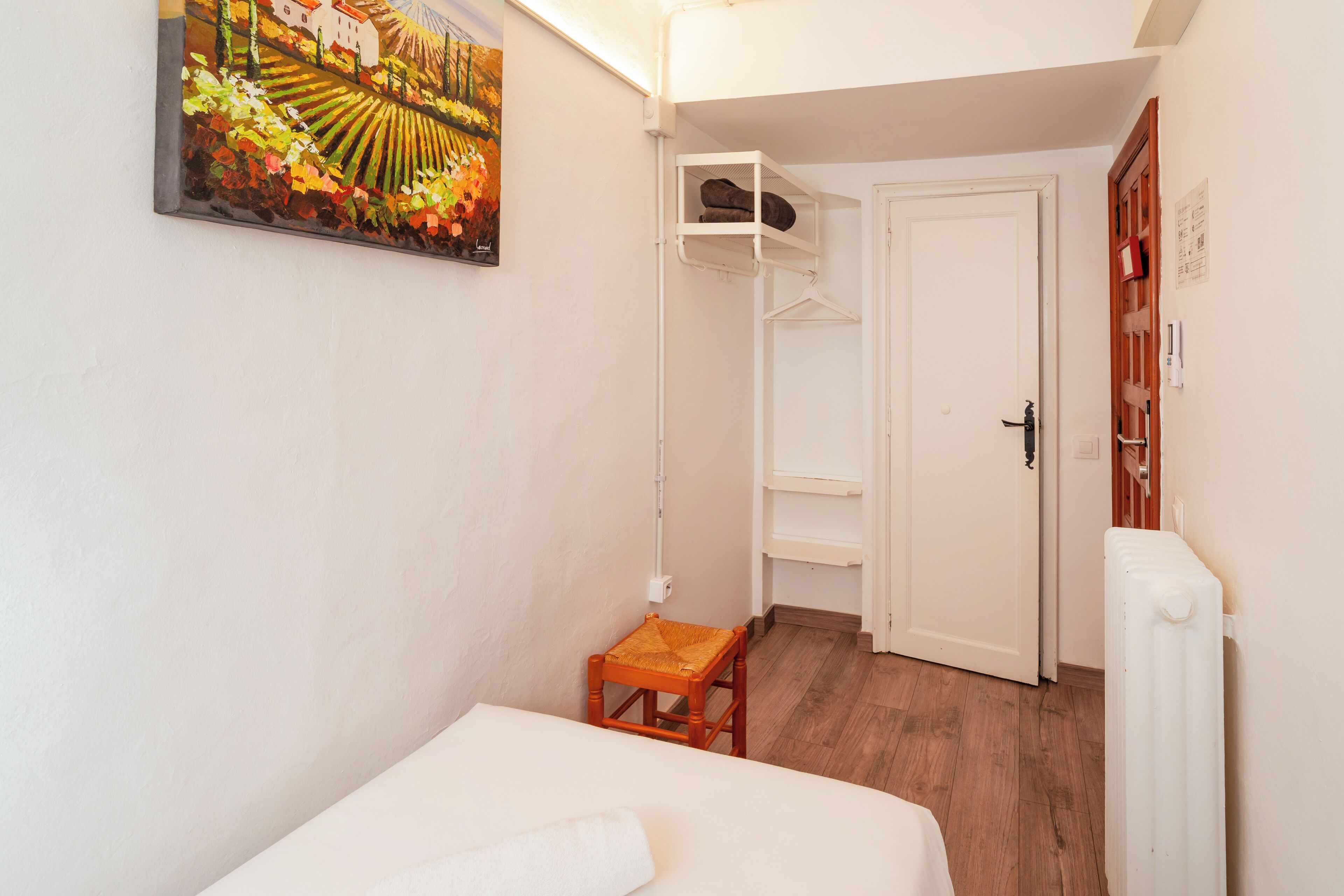 A small, well-lit room with a bed, a wooden chair, a shelf unit, and a radiator.
