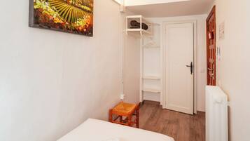 A small, well-lit room with a bed, a wooden chair, a shelf unit, and a radiator.