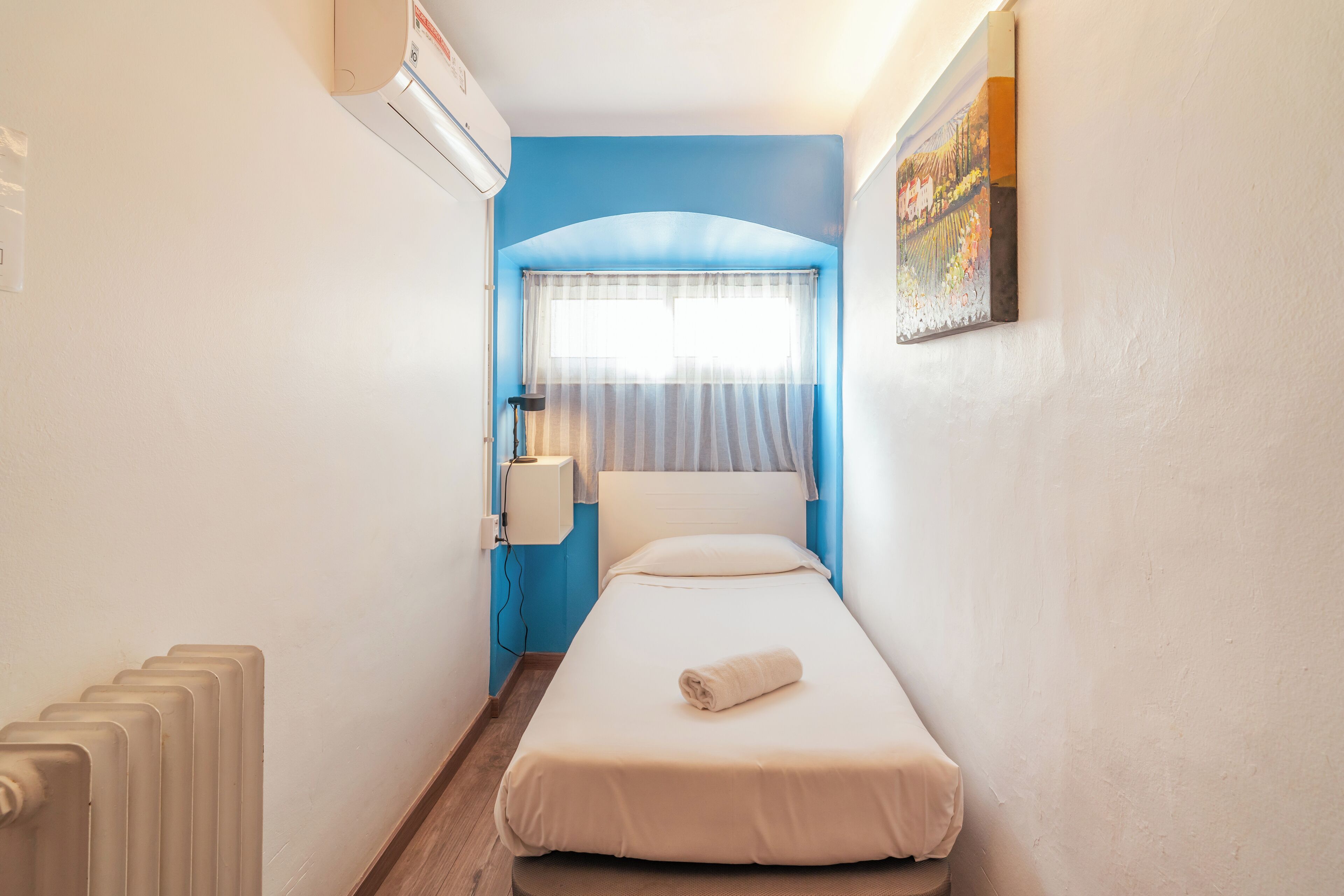 A narrow room with a bed, a radiator, a window with curtains, and a painting on the wall.