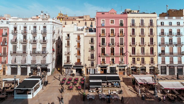 A row of colorful buildings with balconies, a central open square with tables and chairs, and a few storefronts. - Hostal Forum Tarragona (Tarragona)