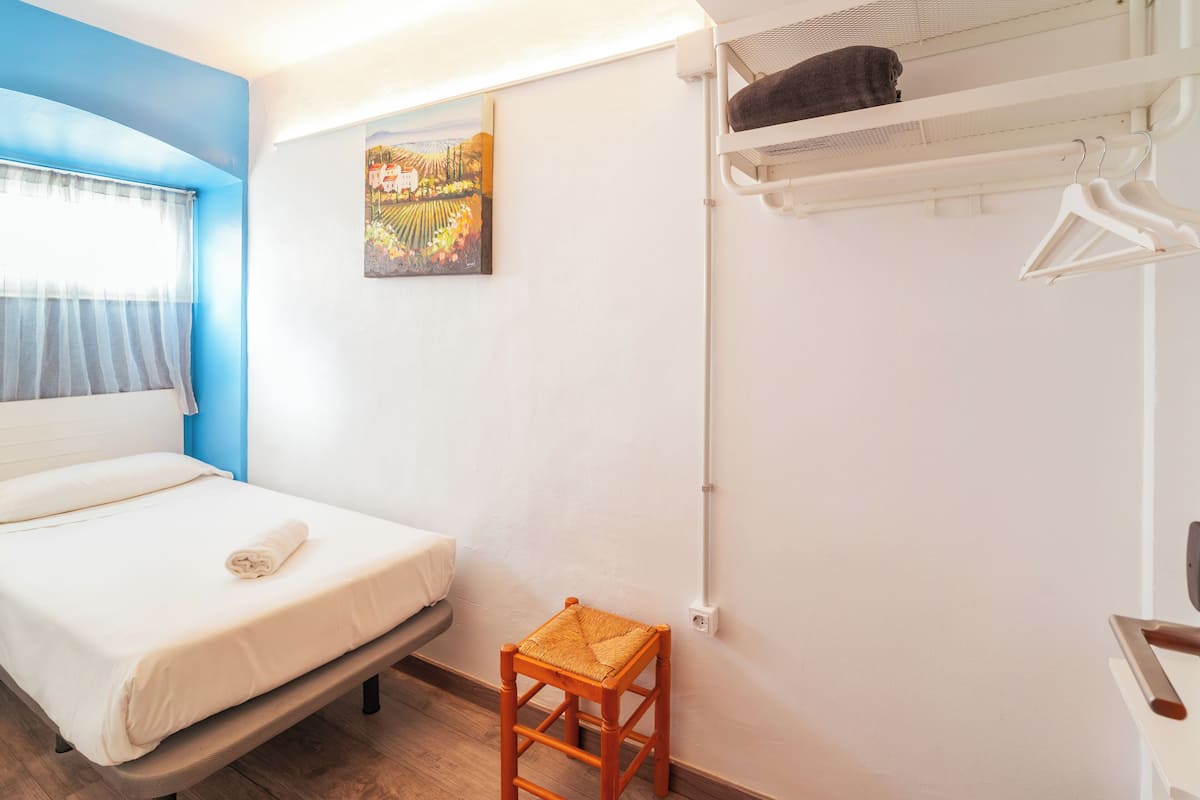 Un dormitorio con cama, un taburete, un cuadro y un tendedero con ropa.