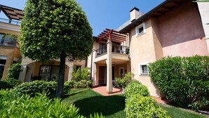 A courtyard with a tree, a pergola, and a building with a balcony. - Appartamento Victoria (Manerba del Garda)