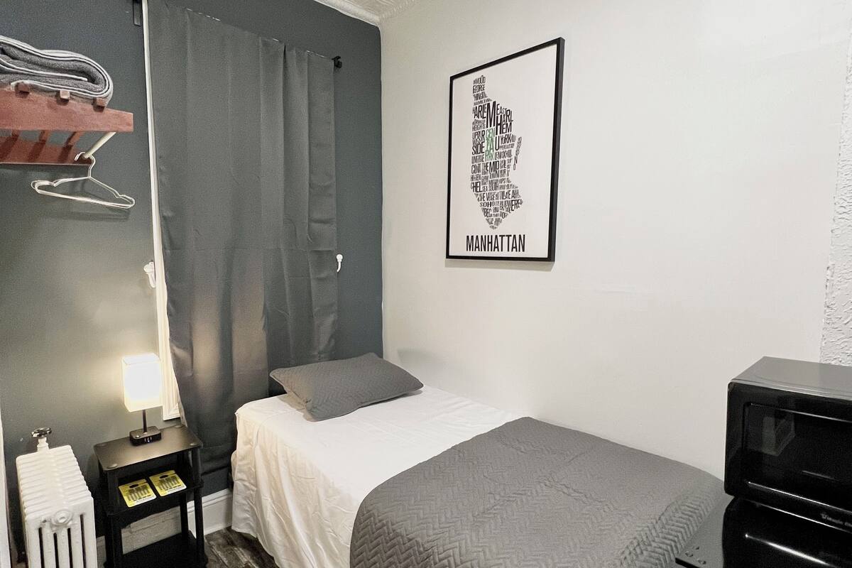Un dormitorio con cama, mesita de noche, radiador, lámpara, manta gris, cómoda negra y un cartel con una nube de palabras en el marco colgado en la pared.
