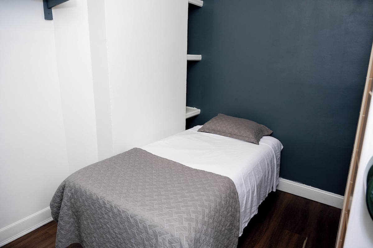 Una cama pequeña y ordenada con colcha gris y ropa de cama blanca en una habitación con paredes azul oscuro y suelo de madera.