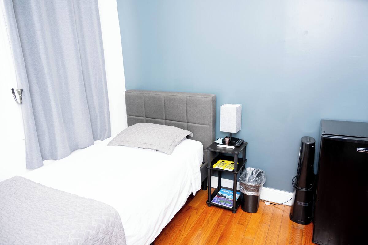 Habitación de hotel con cama, mesita de noche, lámpara, cubo de basura y refrigerador.