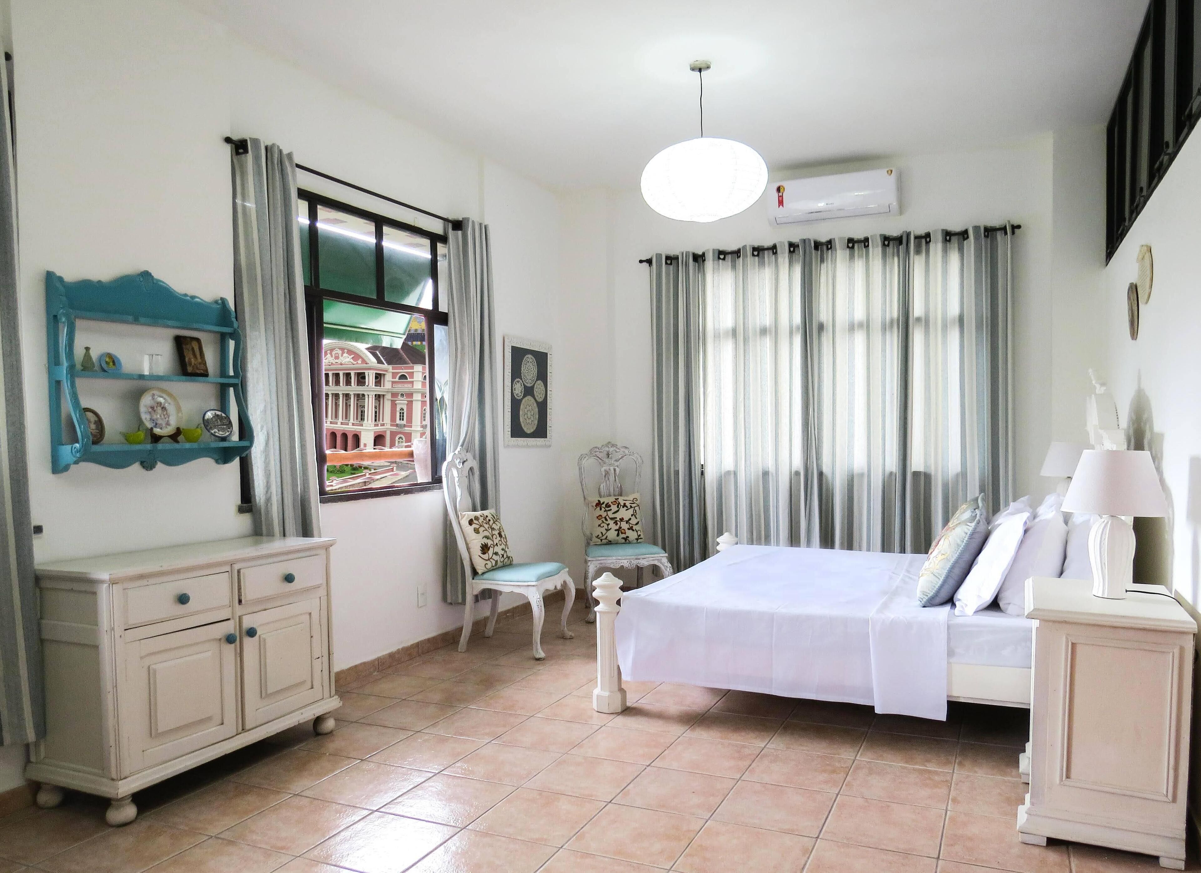 Un dormitorio con cama, cómoda, silla, ventana con cortinas y equipo de aire acondicionado.