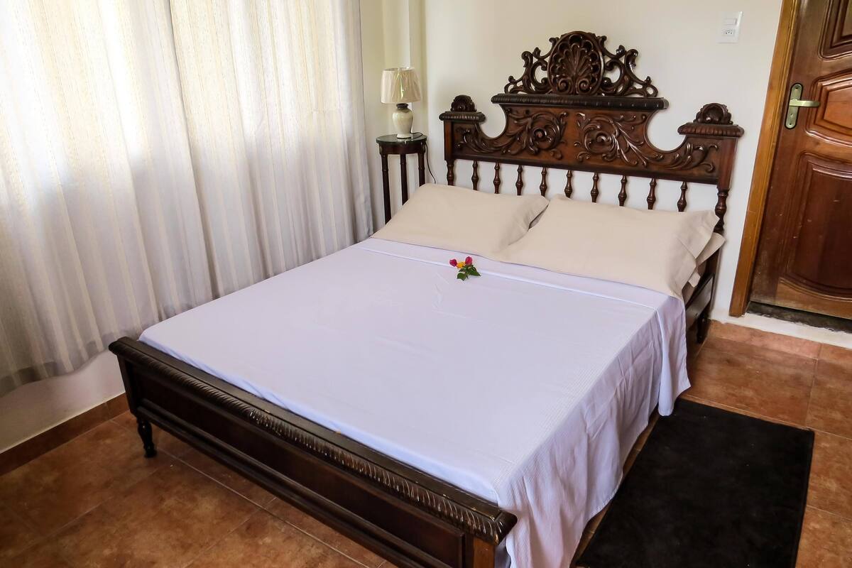 A neatly made bed with a white bedspread and two pillows, a wooden headboard with intricate carvings, a bedside table with a lamp, and a door with a brass handle.