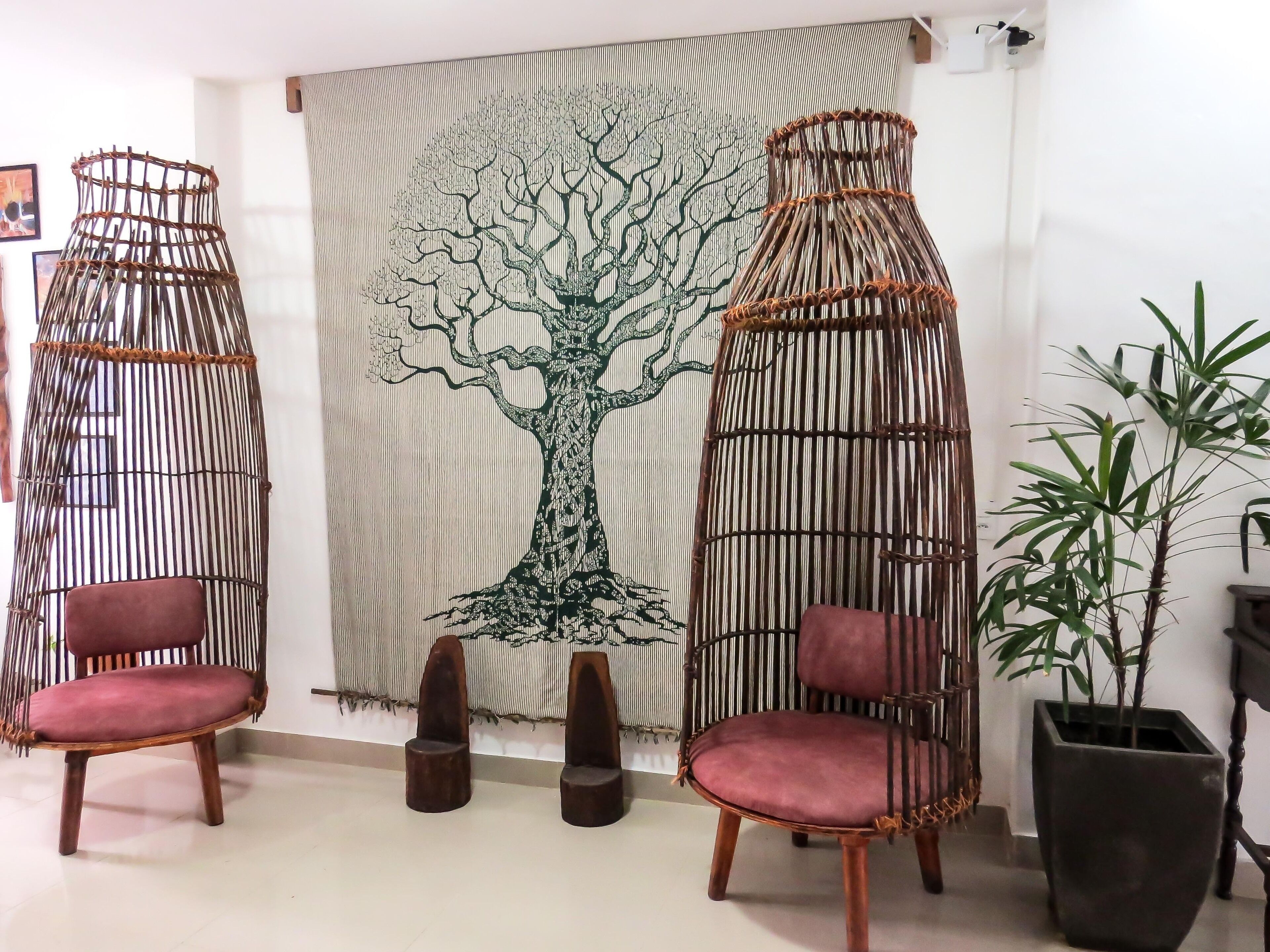 A room with two wicker chairs with cushioned seats, a woven tapestry with a tree design, and a potted plant.