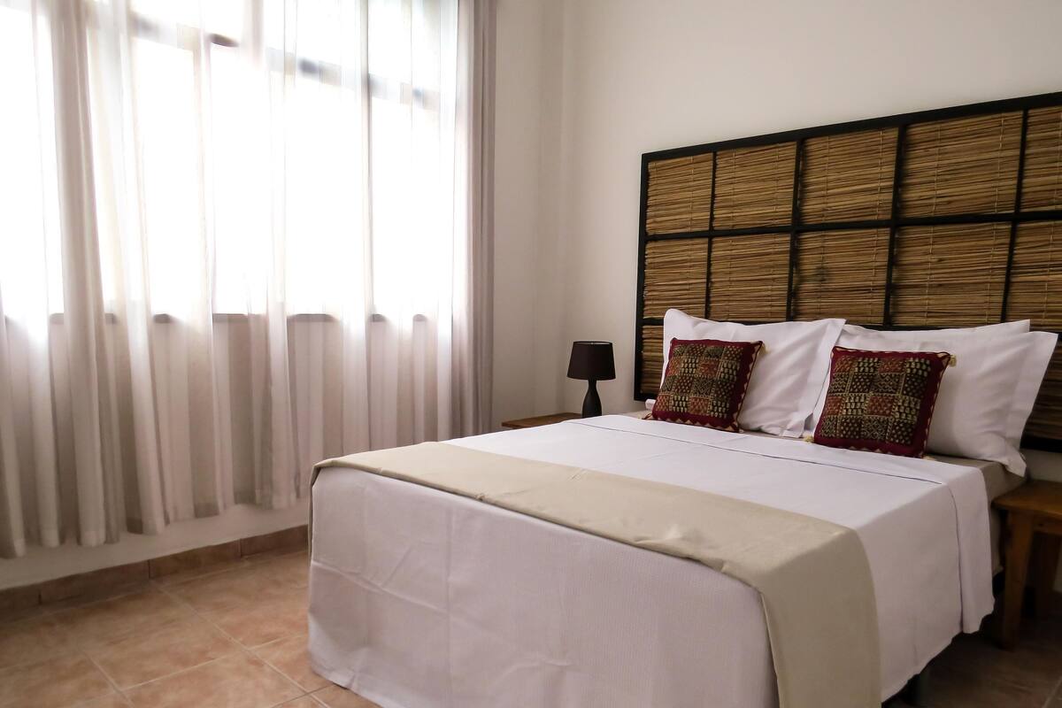A neatly made bed with white linens and two decorative pillows, a bedside table with a lamp, and a window with sheer curtains.