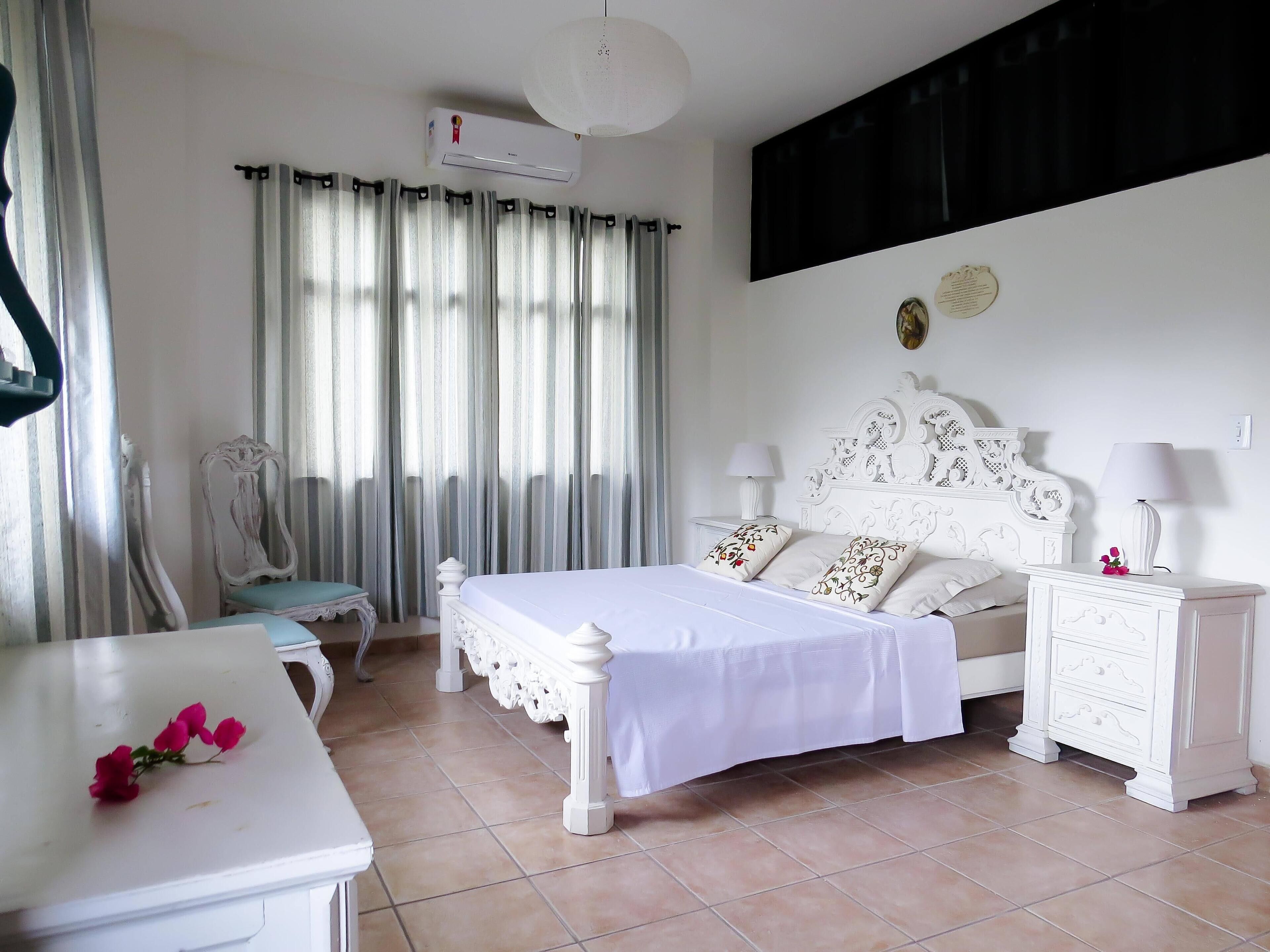 Une chambre à coucher avec un lit blanc, deux tables de chevet, une chaise, un bureau et une fenêtre avec des rideaux.