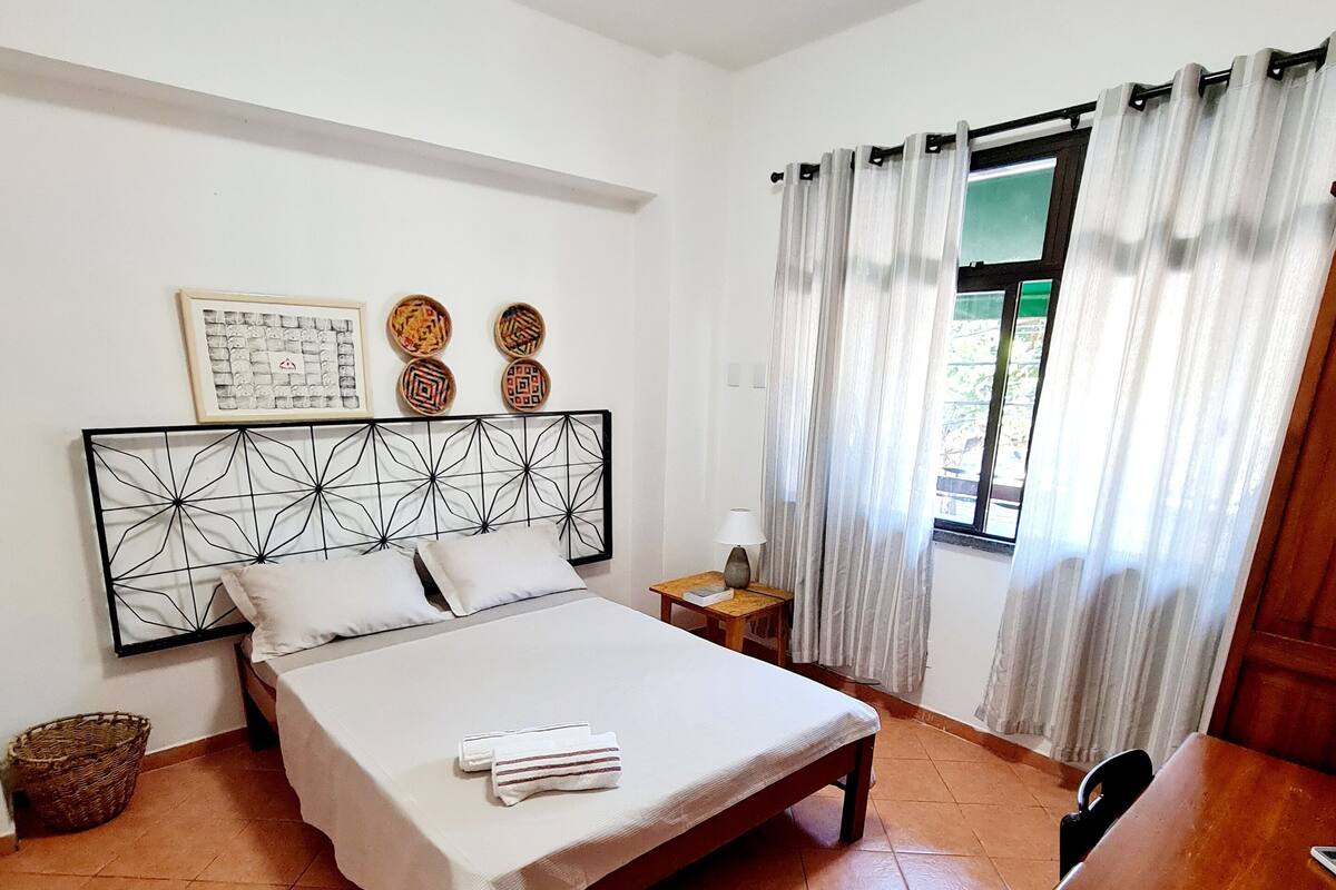 A bedroom with a bed, a nightstand, a lamp, a window with curtains, and a wall with decorative plates.