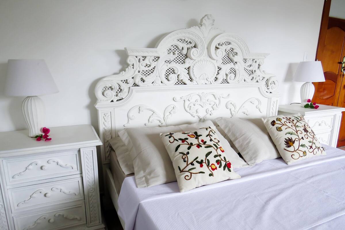 A white bedroom with a decorative headboard, two bedside tables, and a bed with floral pillows.