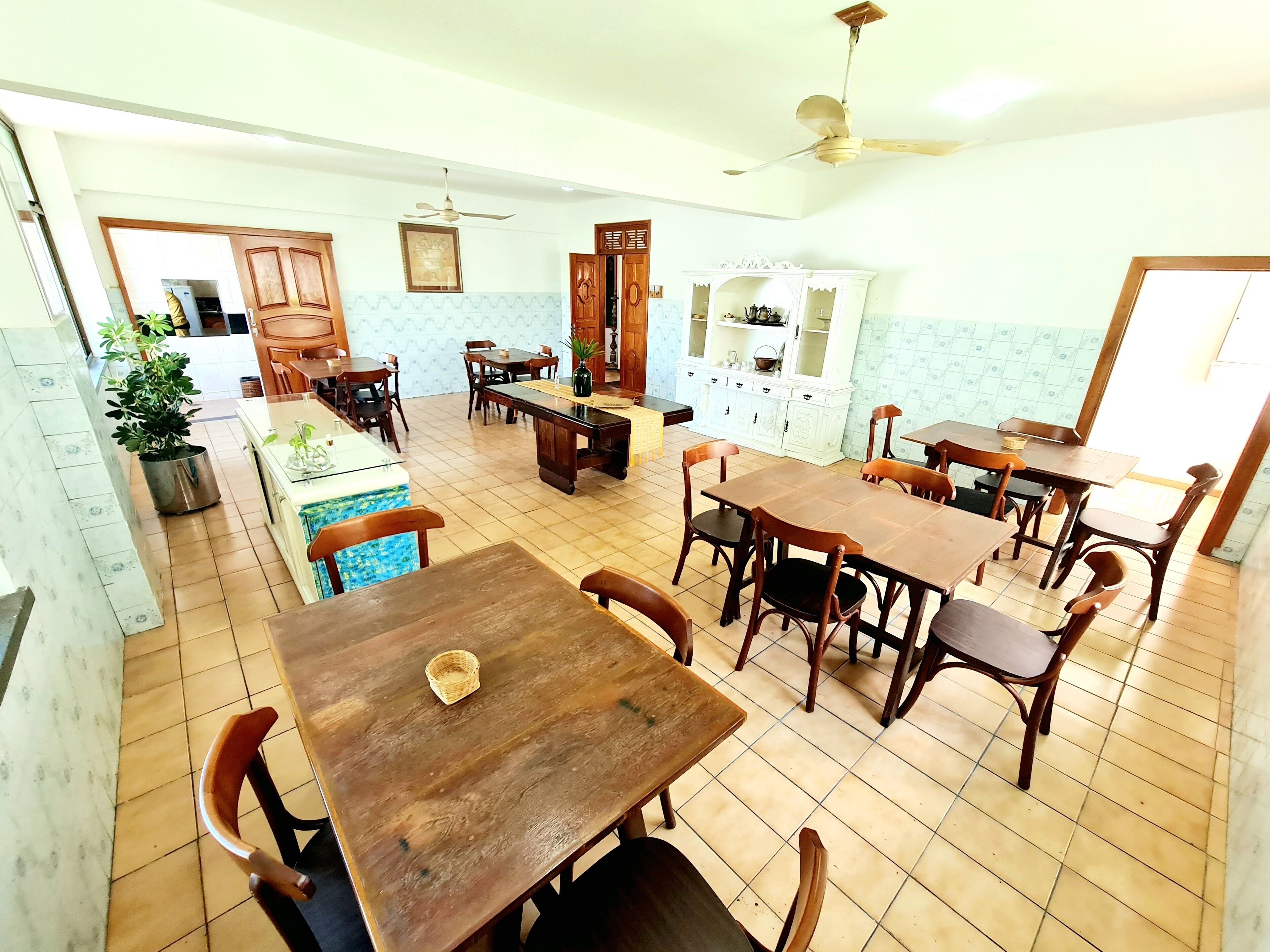 Un comedor amplio con varias mesas y sillas de madera, un ventilador de techo y una zona de cocina con armarios y un lavamanos.
