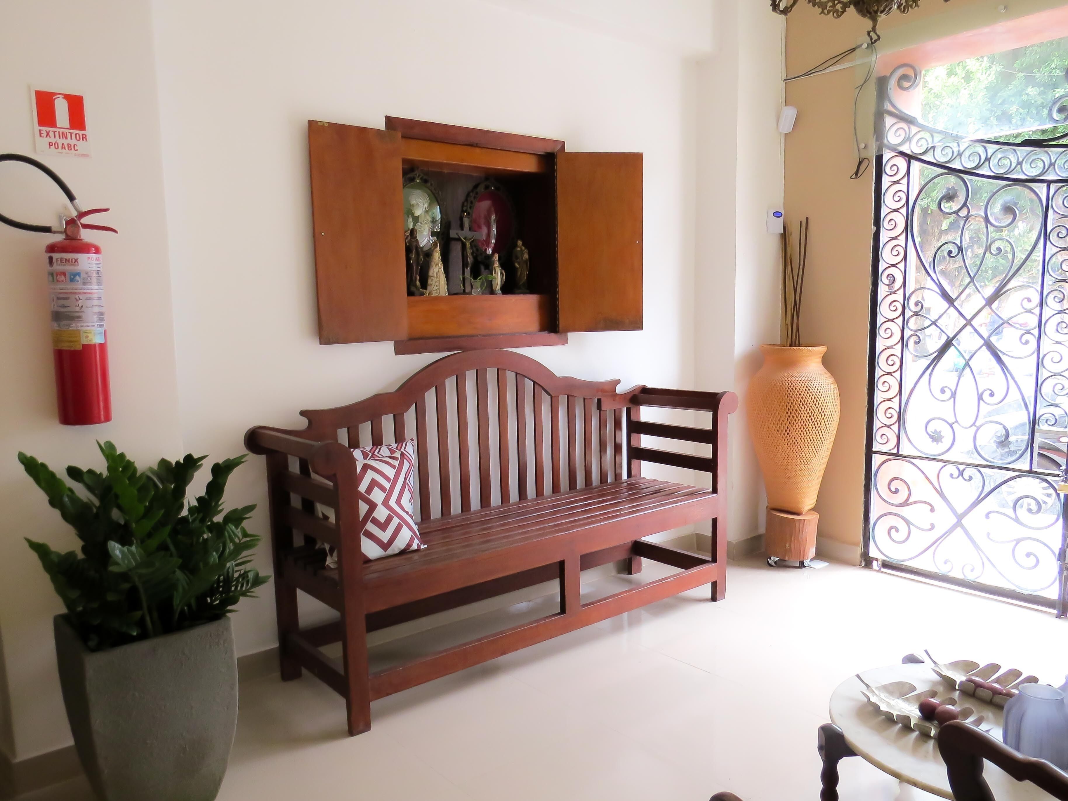 Un banc en bois avec un coussin, une plante en pot, un extincteur et une grille en métal décorative.