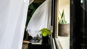 Una habitación con una cama cubierta por cortinas blancas, una mesita de noche con un libro, un florero y una planta en maceta.