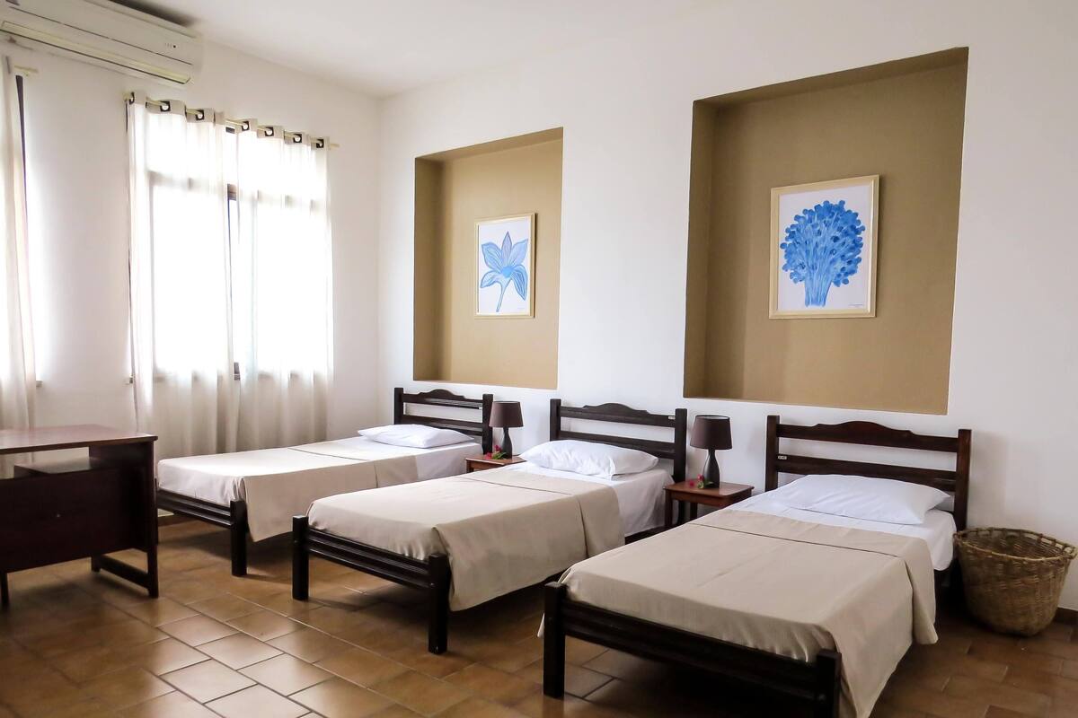 A hotel room with two single beds, a desk, and a chair. There are two framed pictures on the walls, a window with curtains, and a tiled floor.