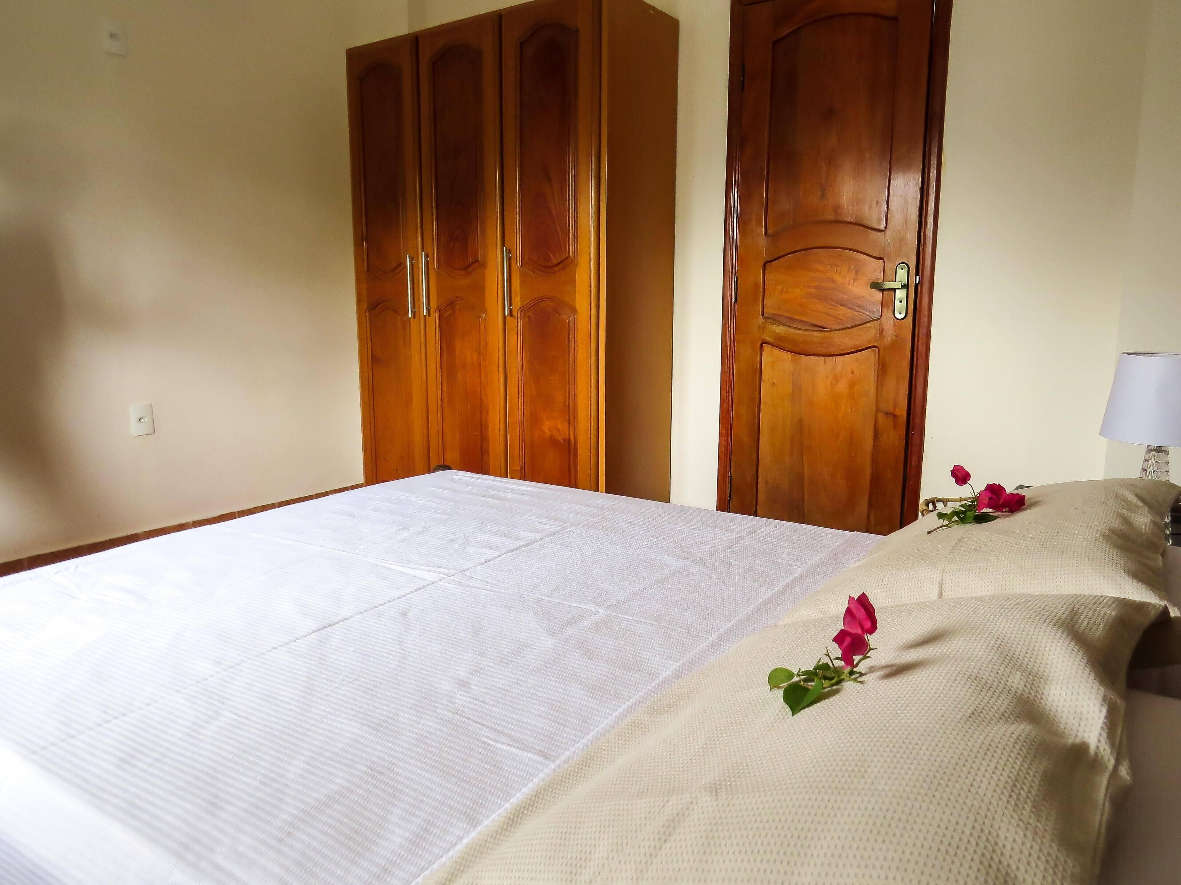 A hotel room with a bed, wooden wardrobe, and a nightstand with a lamp and a rose.