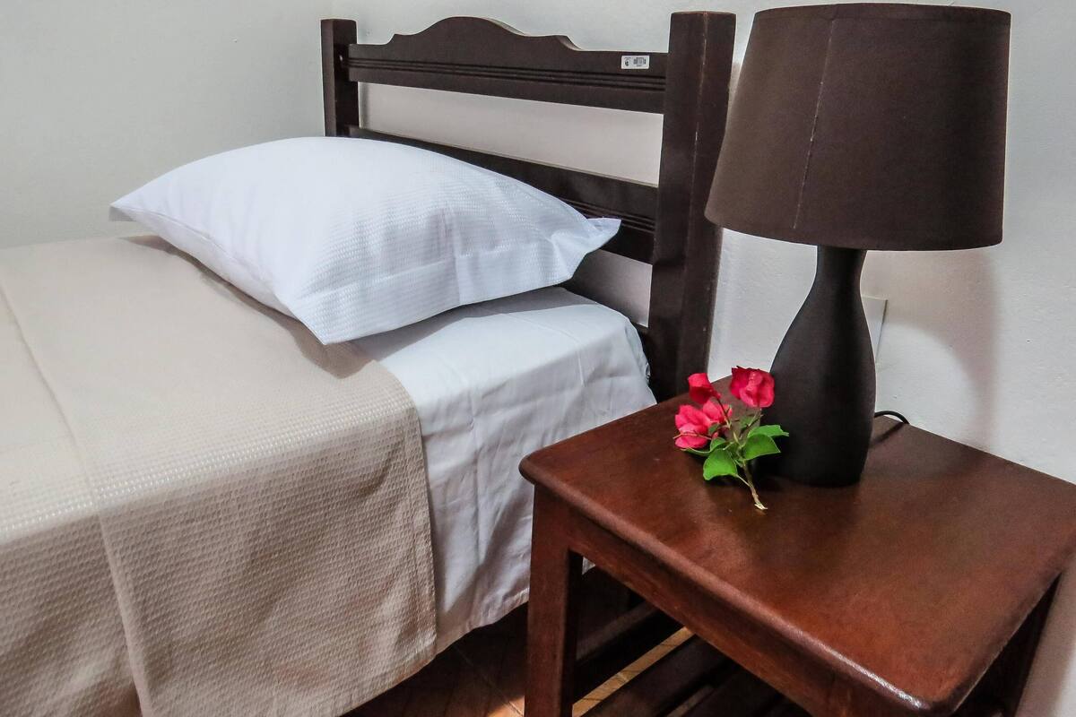 A neatly made bed with a white pillow and a beige bedspread, a dark wooden nightstand with a lamp, and a small table with a vase of red flowers.