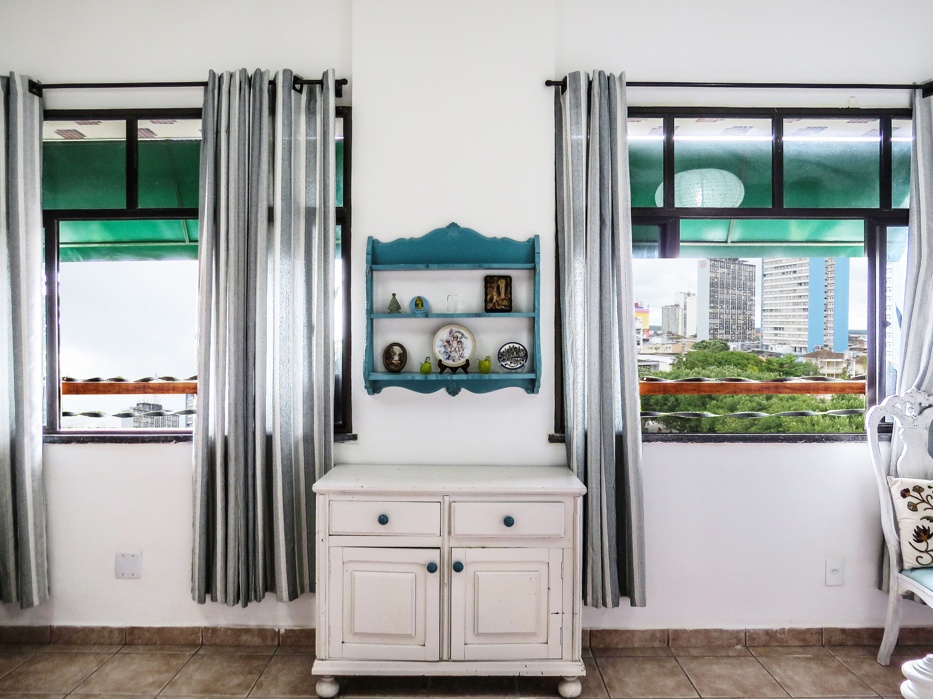 Una habitación con un armario blanco, un estante azul con adornos, y vistas a un paisaje urbano a través de ventanas con tonalidad verdosa.