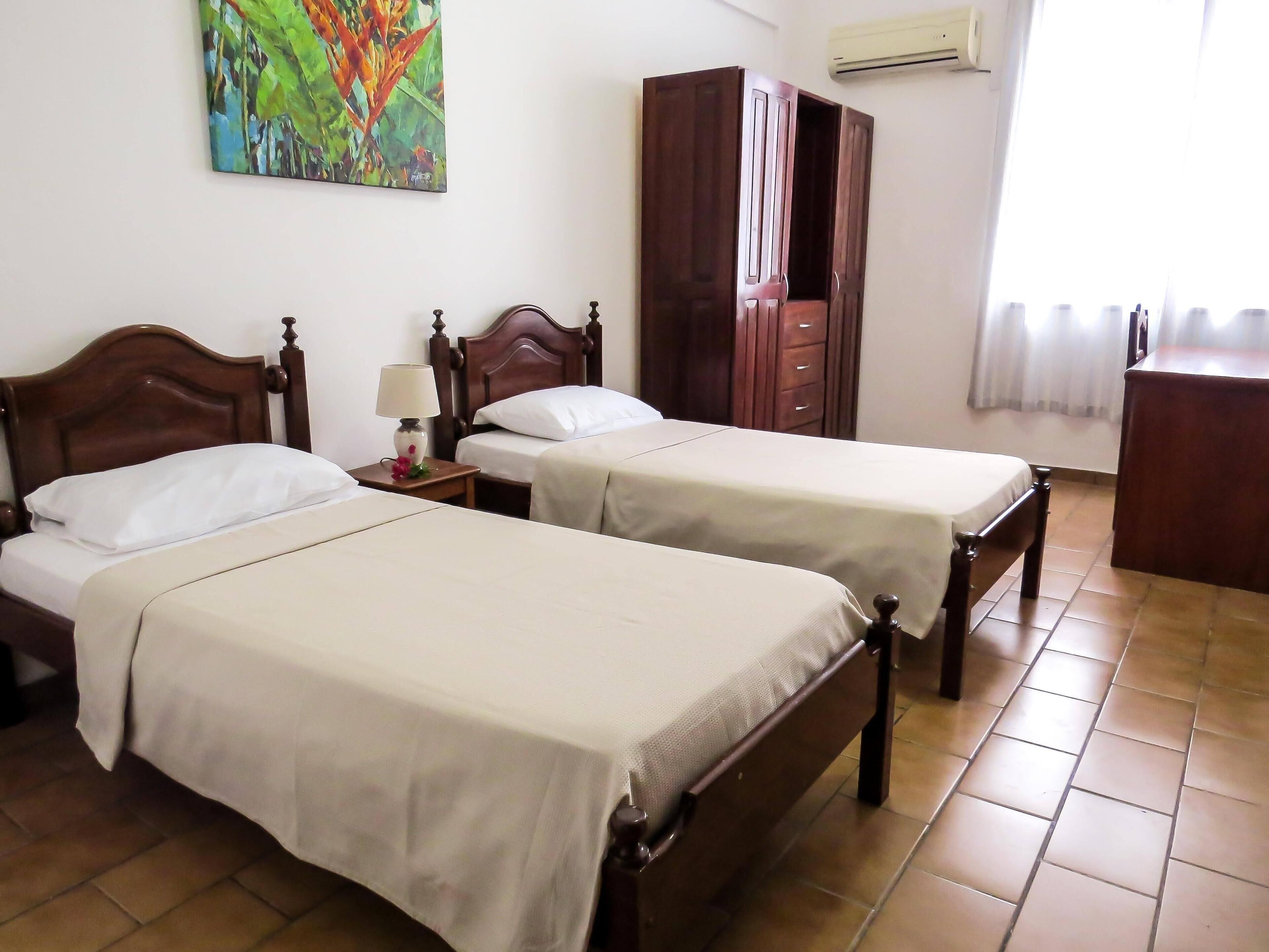A room with two wooden beds, a nightstand with a lamp, a wardrobe, and a painting on the wall.