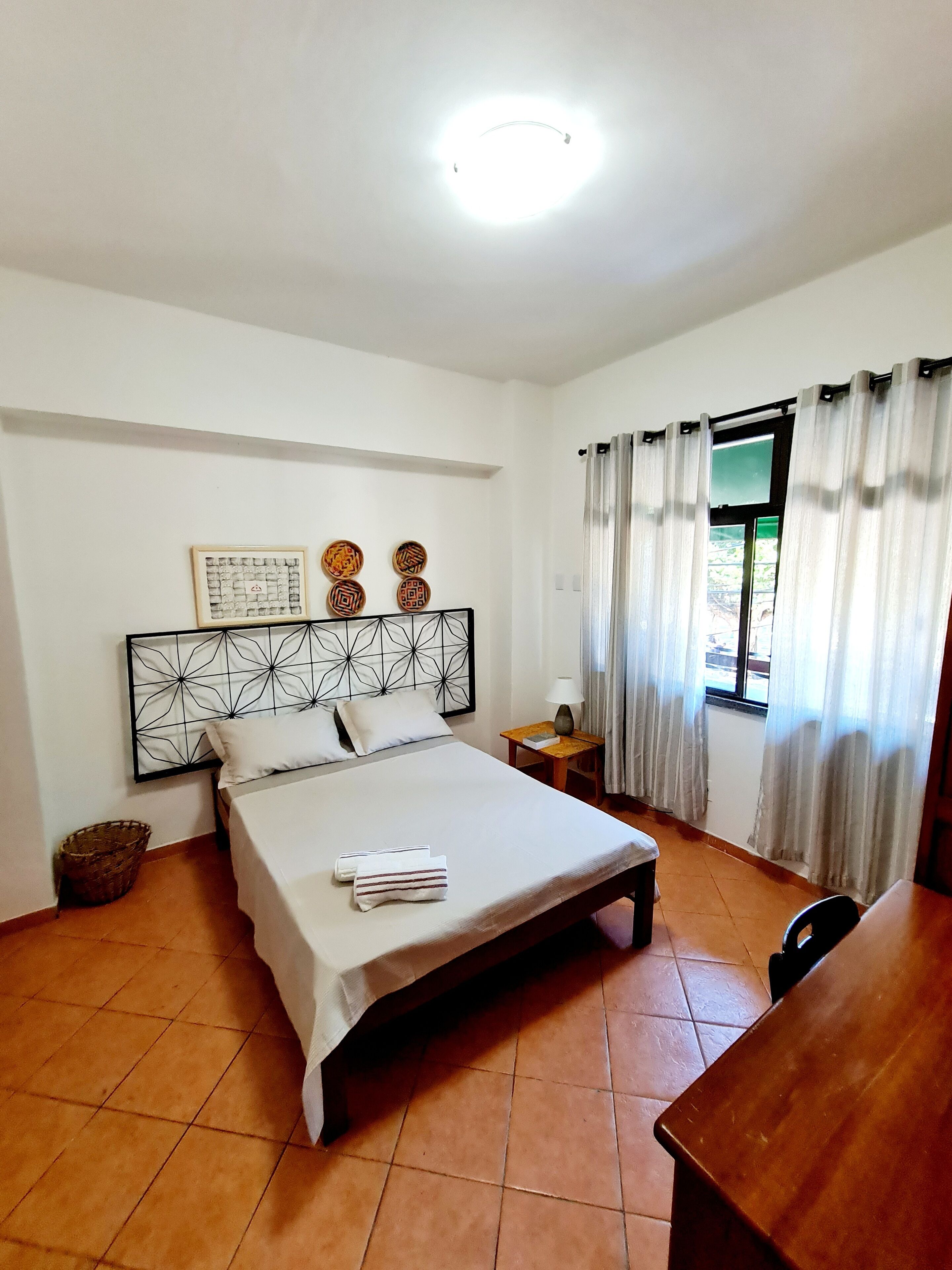 A hotel room with a bed, a bedside table, a lamp, a window with curtains, and a wall with decorative plates.