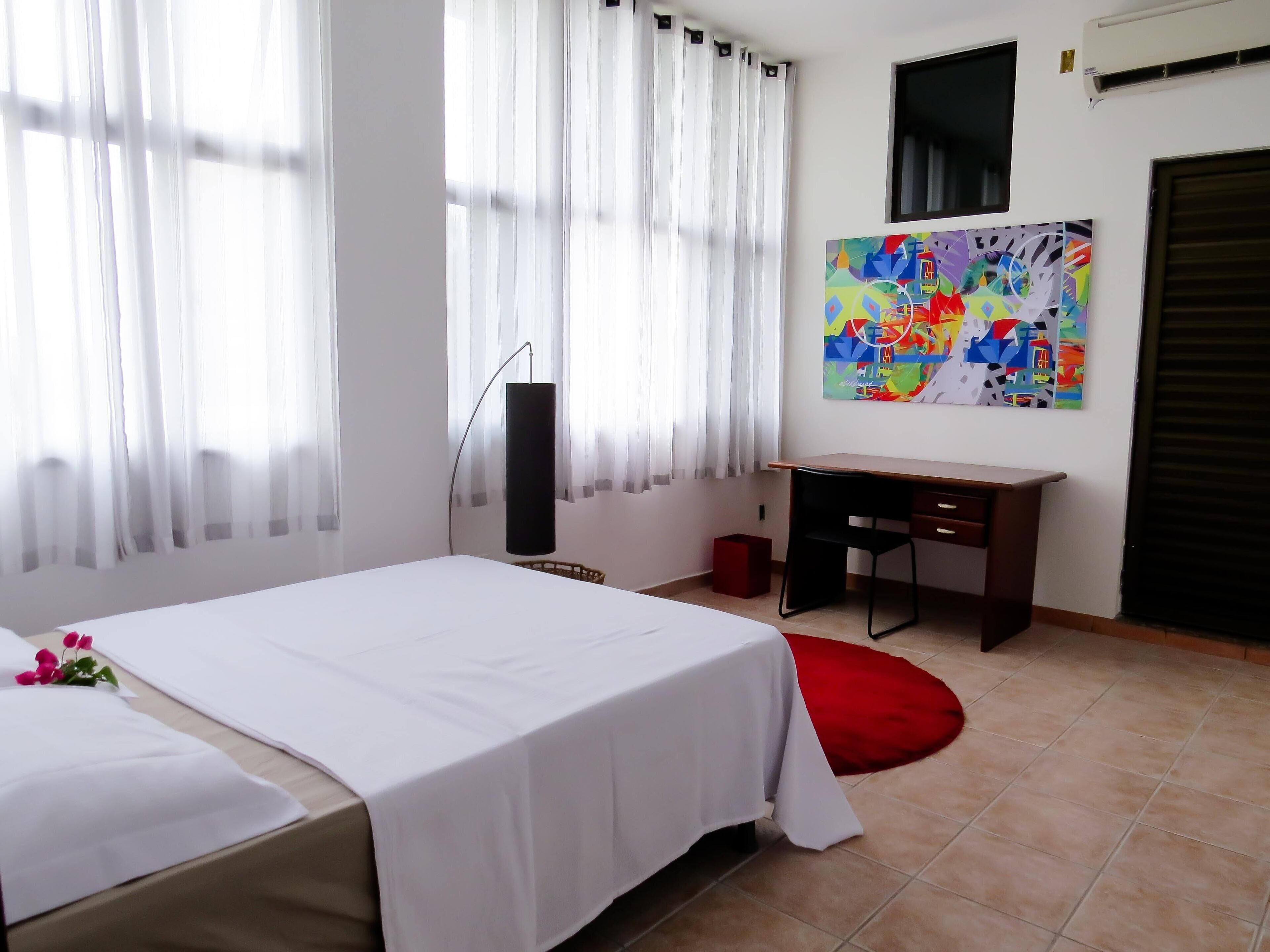 A hotel room with a bed, a desk, a chair, a red rug, and a colorful abstract painting on the wall.