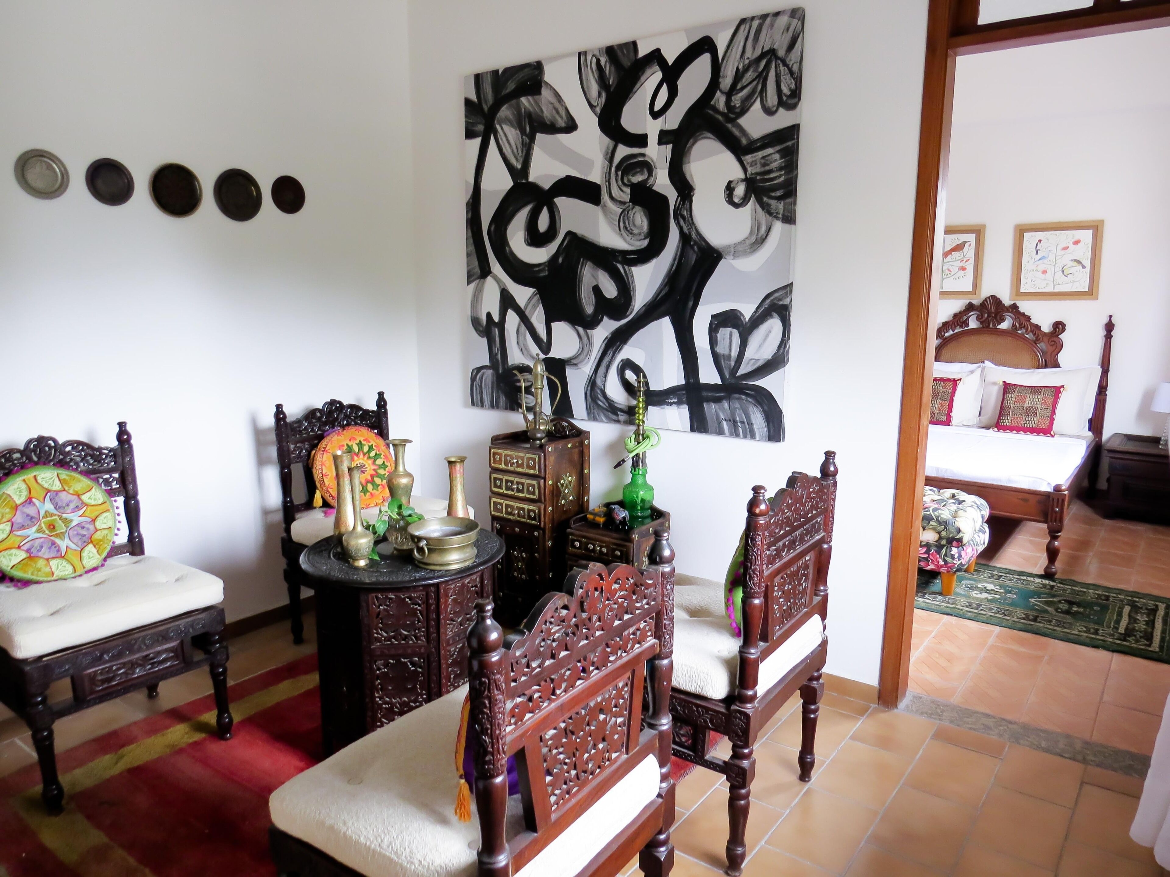 A room with a bed, a wooden chair, a small table with decorative items, and a large abstract painting on the wall.