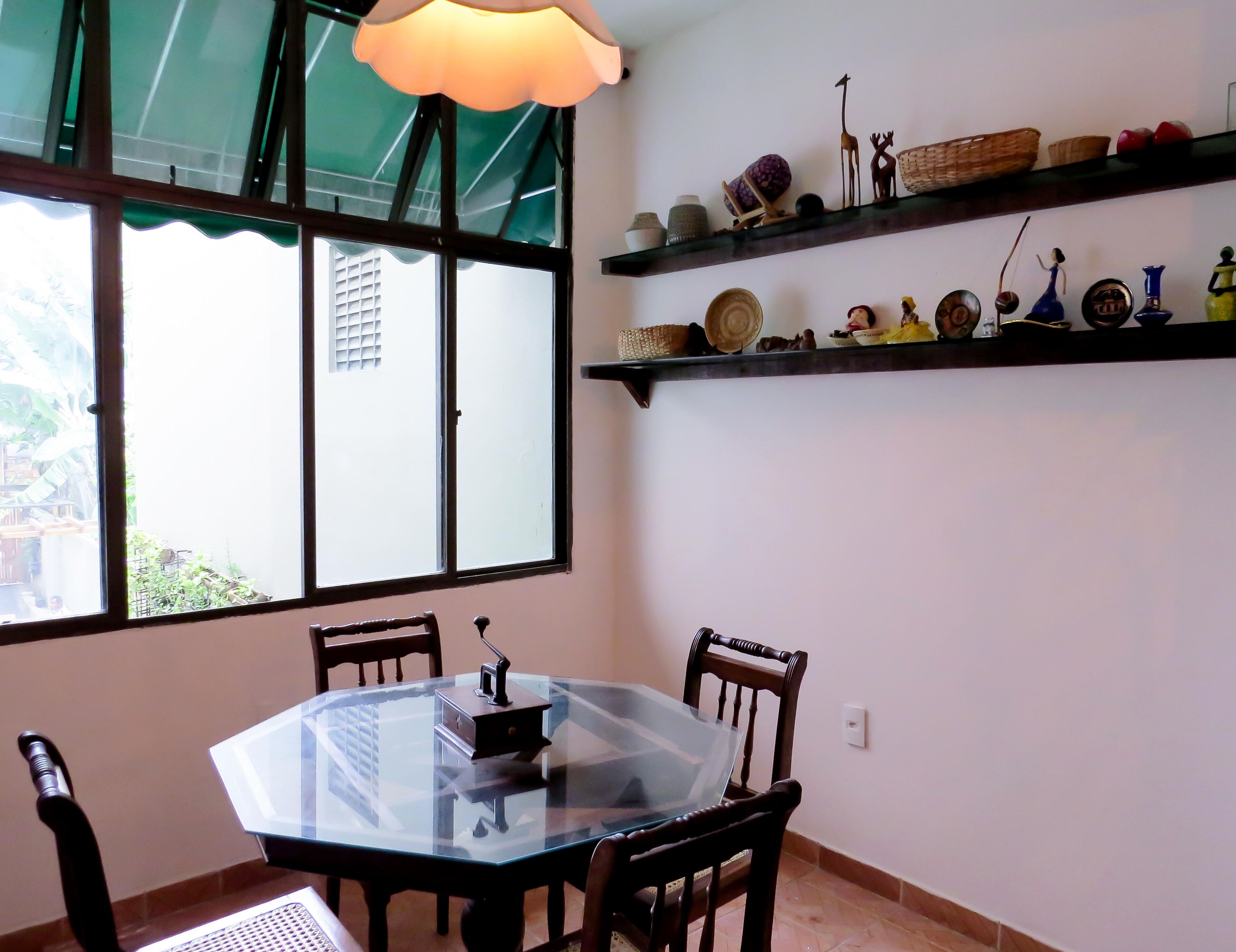 A dining area with a glass-top table and wooden chairs, a window with a green awning, and shelves with decorative items.