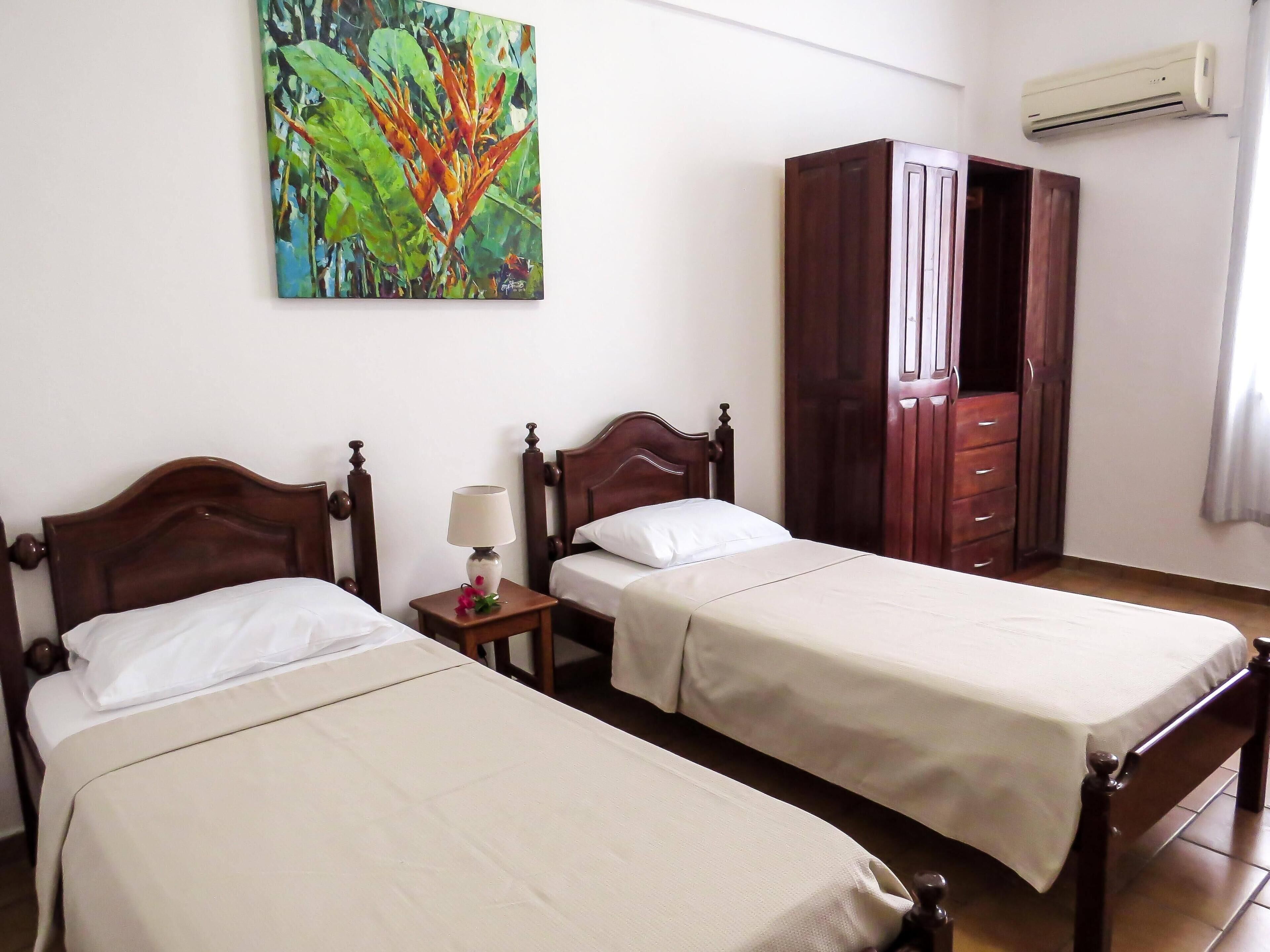 A hotel room with two single beds, a wooden wardrobe, a nightstand with a lamp, and a colorful painting on the wall.
