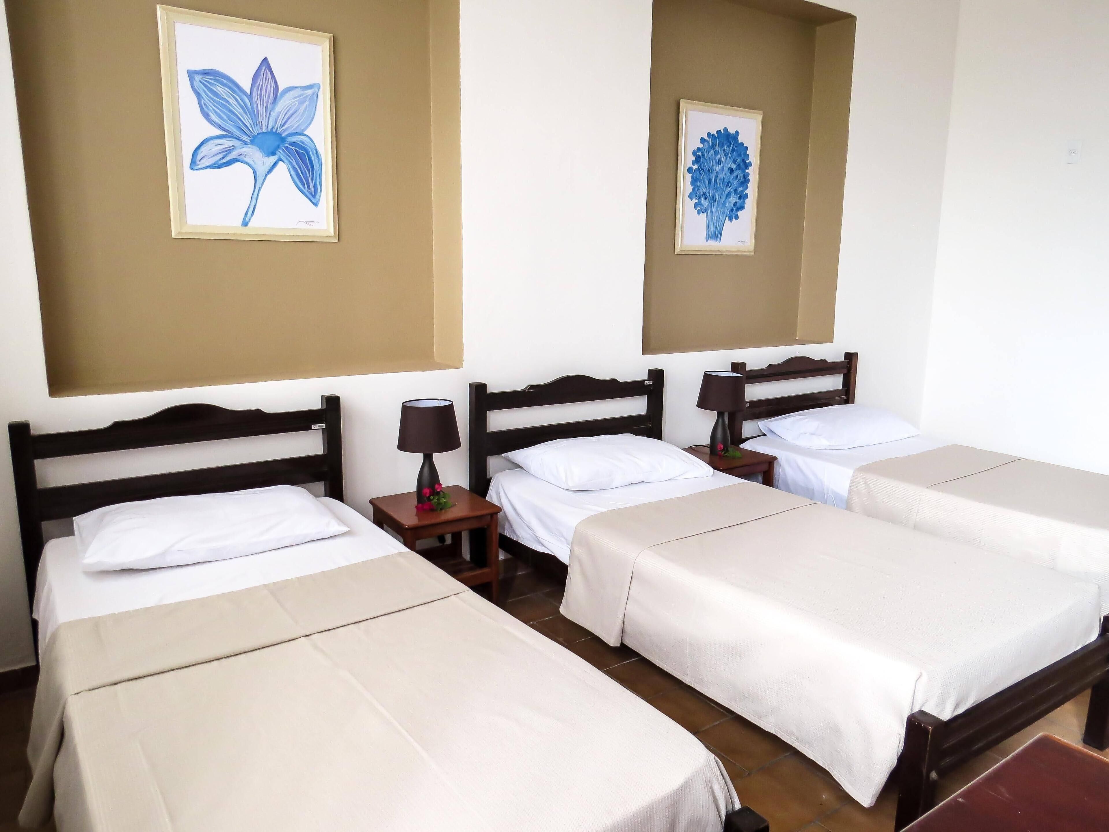 Een hotelkamer met vier eenpersoonsbedden, elk voorzien van een houten hoofdbord, witte lakens en een klein nachtkastje met een lamp.