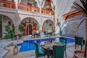 A luxurious courtyard with a swimming pool, multiple dining areas with green upholstered chairs, and ornate architectural details. - Riad Janate & SPA - Suite Familiale (Marrakech)