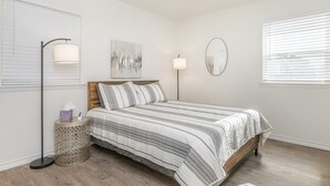 A bedroom with a bed, a ceiling fan, a bedside lamp, a bedside table, and a window with blinds. - 18th Street Retreat  (Dumas)