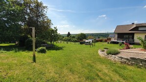 A grassy area with a bench, a stone wall, and a building with solar panels. - Moderne, Top Ausgestattete, Ruhige, Barrierefreie Ferienwohnung (Nonnweiler)
