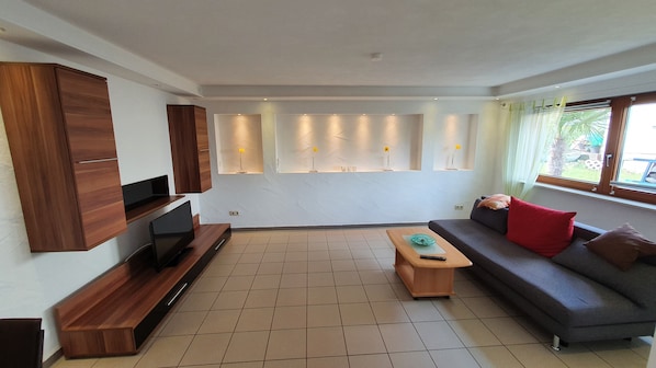A modern living room with a flat-screen TV, a wooden entertainment unit, a grey sofa with red and brown cushions, a wooden coffee table, and recessed lighting on the ceiling. - Moderne, Top Ausgestattete, Ruhige, Barrierefreie Ferienwohnung (Nonnweiler)