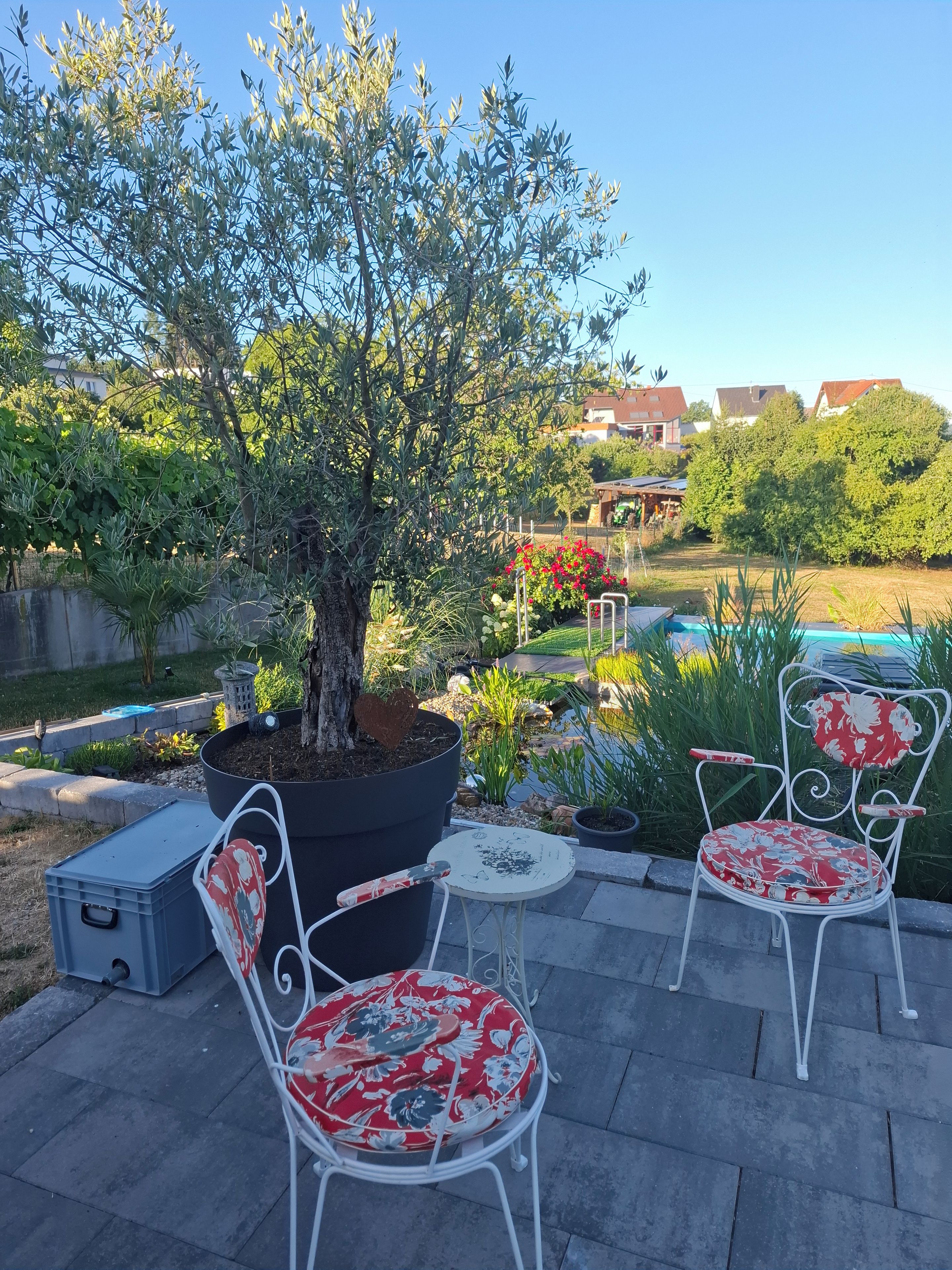 An outdoor seating area with two white metal chairs and a table, surrounded by greenery and a tree.
