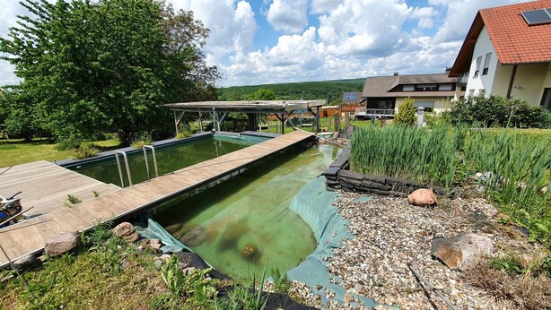 A swimming pool with a wooden deck, surrounded by greenery and a house in the background. - Moderne, Top Ausgestattete, Ruhige, Barrierefreie Ferienwohnung (Nonnweiler)