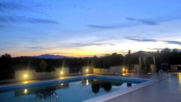 A swimming pool with lights, surrounded by a tiled deck, and a sunset in the background.