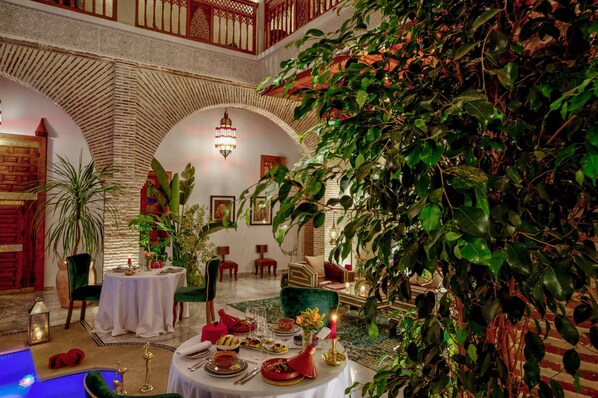 A dining area with a high ceiling, arches, and lush greenery. Tables are set with dinnerware and candles. - Riad Janate & SPA - Chambre Double Standard (Marrakech)
