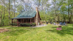 A rustic cabin with a stone chimney, surrounded by a grassy yard and trees. - Creekside, Hot Tub, Fire Pit, Fireplace, Quiet, Easy Access (Cosby)