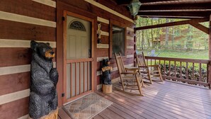 A wooden cabin with a porch, a welcome sign, a bear sculpture, and a view of the surrounding forest. - Creekside, Hot Tub, Fire Pit, Fireplace, Quiet, Easy Access (Cosby)