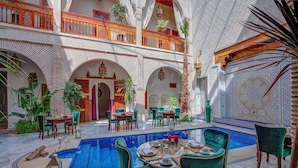 A luxurious courtyard with a swimming pool, multiple dining areas with green upholstered chairs, and ornate architectural details. - Riad Janate & SPA (Marrakech)