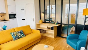 Un salon moderne avec un canapé jaune, un fauteuil bleu, une table basse en bois et une vue sur la mer grâce à de grandes fenêtres.