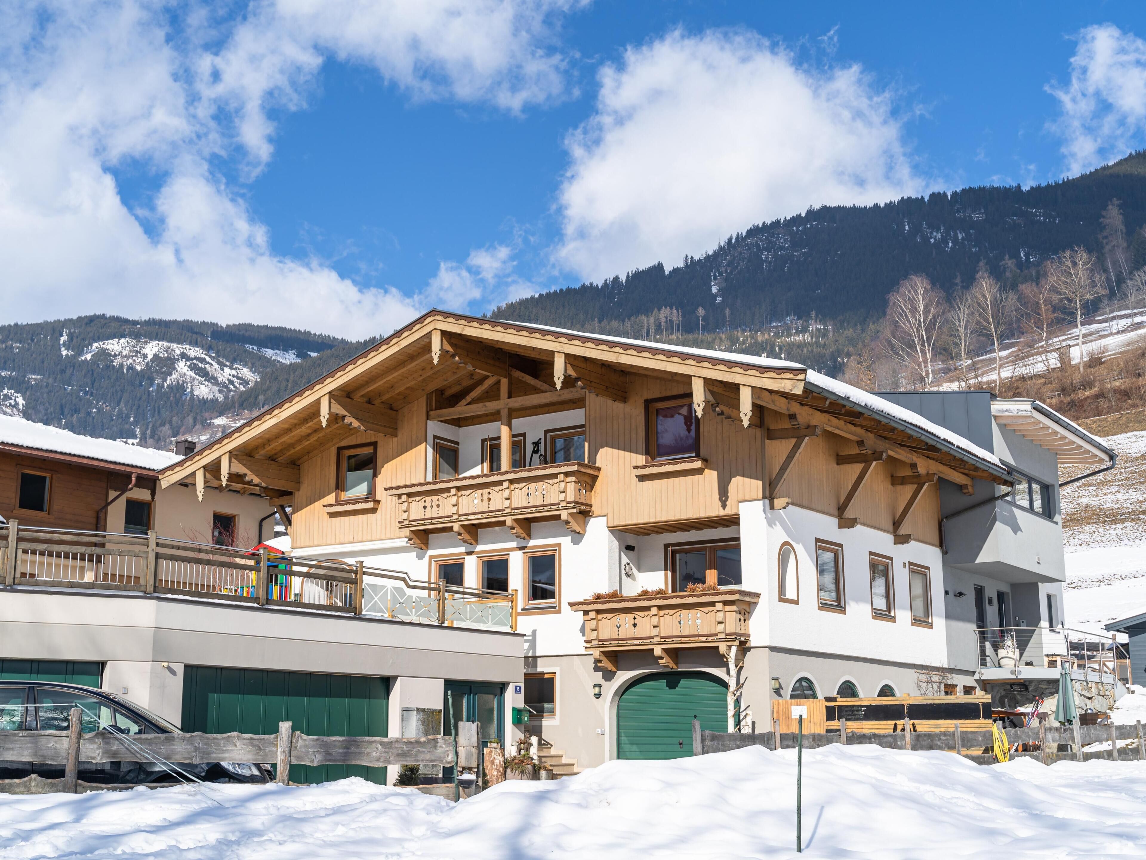 A two-story wooden chalet with a balcony, a green garage door, and a snowy mountain backdrop.