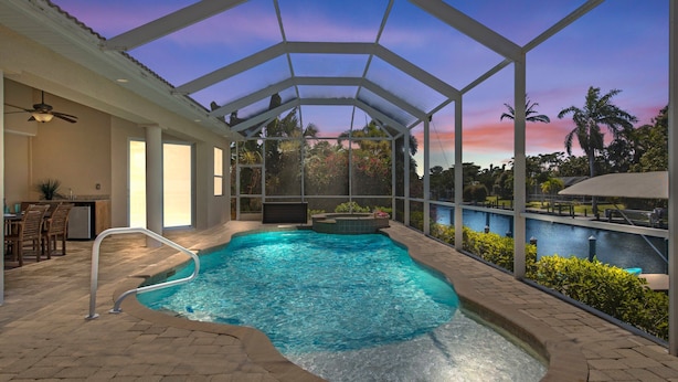A covered swimming pool with a glass ceiling, a hot tub, and a view of a body of water and palm trees. - WALK TO BEACH! - Luxury Waterfront w/4 ensuites, Pool/Spa, Bikes, Kayaks, Dock/ Direct Gulf Access (Cape Coral)