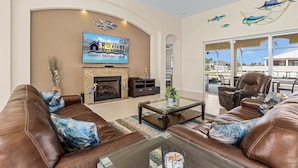A spacious living room with leather sofas, a glass coffee table, a fireplace, and a TV mounted on the wall. - WALK TO BEACH! - Luxury Waterfront w/4 ensuites, Pool/Spa, Bikes, Kayaks, Dock/ Direct Gulf Access (Cape Coral)