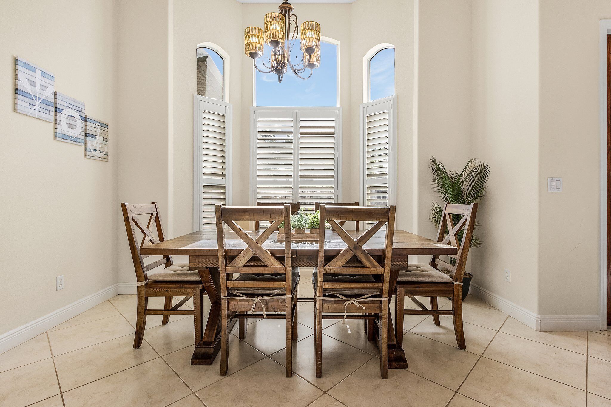 Un comedor con mesa y sillas de madera, una araña y vistas a través de contraventanas blancas.