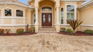 A two-story house with a beige facade, a brown double door with glass panels, and a tiled roof. - WALK TO BEACH! - Luxury Waterfront w/4 ensuites, Pool/Spa, Bikes, Kayaks, Dock/ Direct Gulf Access (Cape Coral)